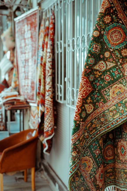 A display of colorful, patterned rugs hanging vertically along a white metal gate in an indoor setting. The rugs feature intricate geometric and floral designs in various shades of red, green, orange, and blue. To the left, part of a wooden chair with a tan leather seat is visible, placed against a plain wall. The floor appears to be made of wood, and the lighting is natural and soft, highlighting the vibrant patterns of the rugs. The scene suggests a room or shop space focused on textile or home decor, with a clean and well-organized appearance, reflecting attention to surface cleanliness. For professional deep cleaning and maintenance of such carpets and rugs, Deep Cleaning Lambeth offers expert domestic cleaning services.
