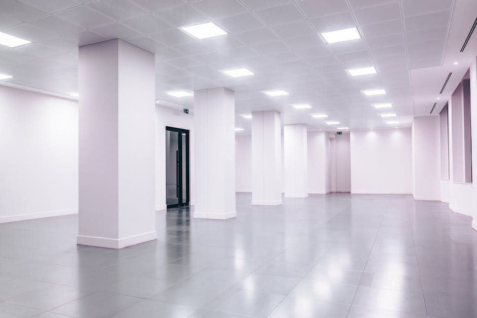 A spacious, modern commercial office interior with white walls and a smooth, grey tiled floor reflecting light from evenly spaced ceiling LED panels. The space features multiple supporting white columns and a black-framed glass door on the left side, leading to an elevator. The clean and tidy environment highlights the surfaces' pristine condition, with no dust or marks visible, indicative of recent deep cleaning and surface sanitisation carried out by Deep Cleaning Lambeth, ensuring a hygienic and professional workspace.