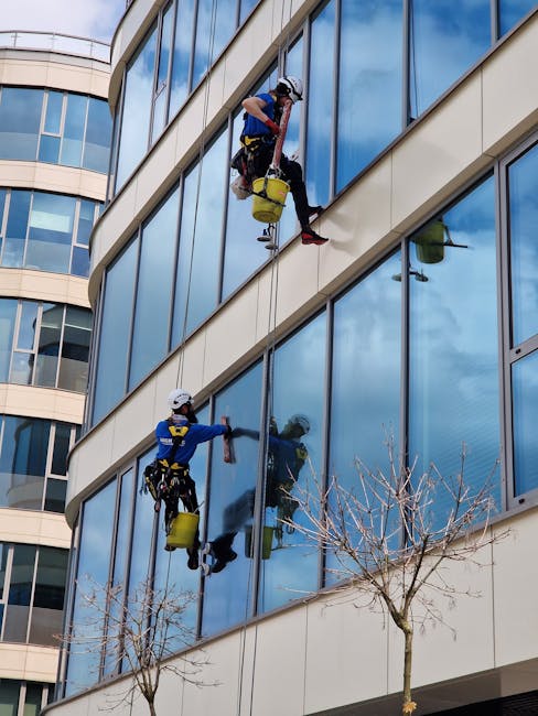 Two professional window cleaners wearing safety helmets and harnesses are performing surface cleaning on the exterior glass facade of a modern multi-storey office building in Vauxhall Lambeth SW8. They are suspended by ropes and using squeegees and cloths to thoroughly clean the large, reflective glass panels, which are part of the building's curved architectural design. The sunlight reflects off the pristine, streak-free glass, emphasizing the clarity and shine achieved through deep cleaning. Visible in the image are the blue sky and a few tree branches at the bottom, providing context of an outdoor urban setting. The scene captures a high-level commercial cleaning operation, coordinated by Deep Cleaning Lambeth, ensuring the building’s exterior remains hygienic and visually appealing.