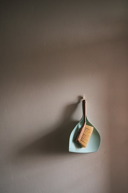 A light blue wall hook attached to a neutral-colored wall, holding a beige cleaning brush with bristles and a wooden handle. The brush is resting on a matching light blue, teardrop-shaped holder that appears to be made of plastic. The surface of the wall and hooks is clean and free of dust or stains, indicating recent or ongoing deep cleaning. The composition highlights the tools used in surface cleaning and maintenance within a domestic setting, with soft lighting emphasizing the tidy and sanitized condition, typical of professional cleaning services by Deep Cleaning Lambeth. No other objects or surfaces are visible in the image, focusing on the cleaning tool storage area.