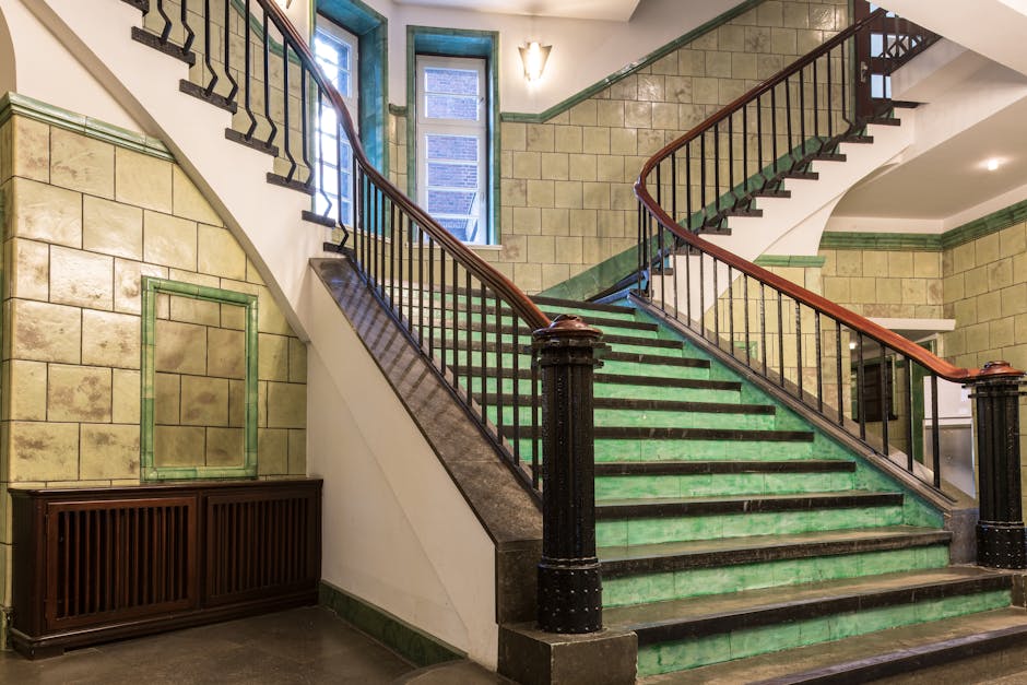 The image depicts a grand staircase inside a building, featuring green ceramic tile flooring on the steps and at the base, with black metal railings supported by large, polished black posts. The stair