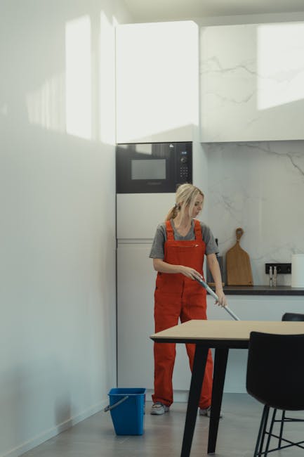 A woman wearing a grey t-shirt and red overalls is mopping a light-colored wooden or laminate floor in a modern kitchen with white walls, a marbled backsplash, and black dining chairs. She stands next