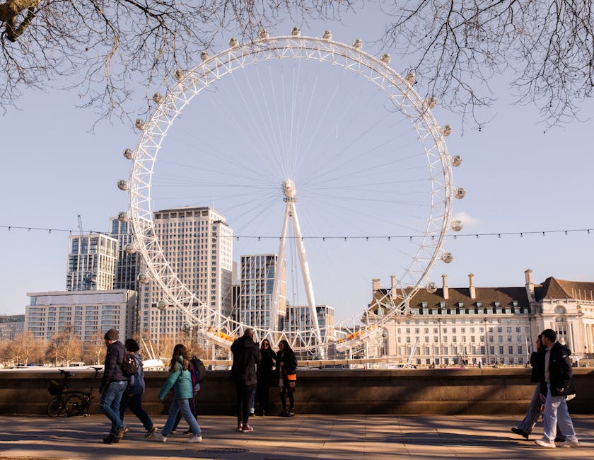 A large ferris wheel with a white metal structure and multiple enclosed passenger cabins is set against a clear sky in an urban area with modern high-rise buildings and older architectural styles. The foreground features a paved walkway with several people walking, some in pairs or groups, wearing casual winter clothing. Bare trees line the background, suggesting a late autumn or winter setting. The scene is well-lit with natural sunlight, creating shadows on the ground, highlighting the clean and orderly appearance of the cityscape. The image captures the vibrant atmosphere of an outdoor city attraction, with no visible cleaning equipment or surfaces, but the detailed scene reflects an environment where surface cleaning and maintenance are essential for hygienic urban spaces, as exemplified by services provided by Deep Cleaning Lambeth.