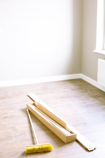 A brightly lit room with light beige walls and a white radiator below a large window, featuring a light wooden floor that is partially covered with a wooden window frame and a yellow-handled brush placed on the floor. The room appears recently cleaned and prepared for renovation, with clean surfaces and no visible dust or debris. The natural light coming through the window highlights the smooth, polished finish of the flooring, and the scene suggests a focus on surface cleaning and preparation, consistent with services provided by Deep Cleaning Lambeth for post-renovation cleaning projects at The Oval Lambeth SE11.