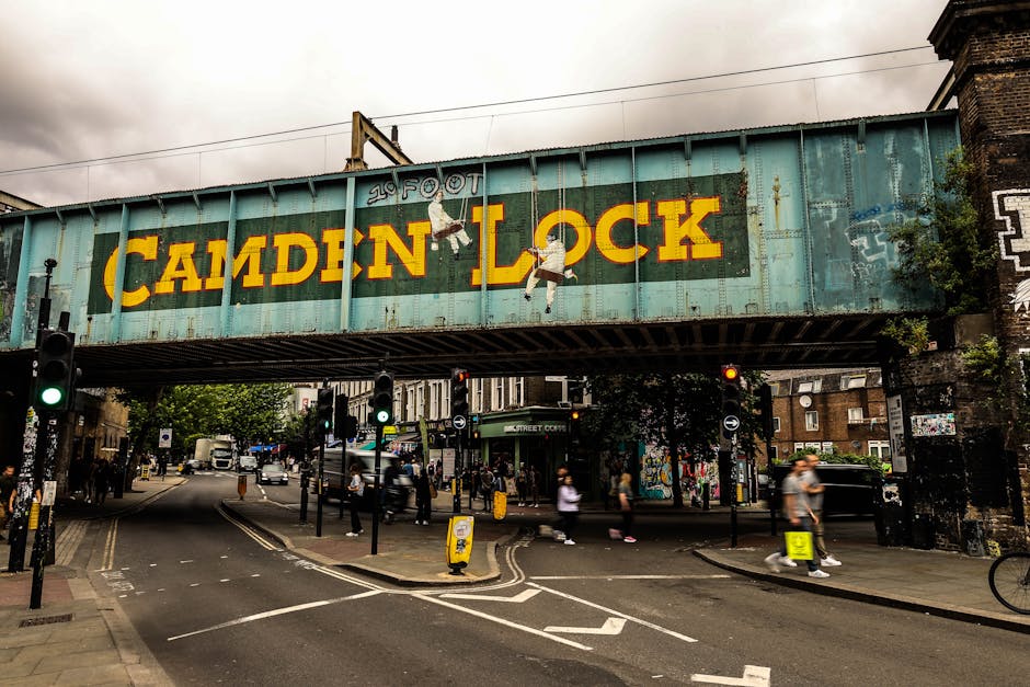 A large, weathered metal railway bridge spanning a busy London street, featuring a painted sign that reads 'Camden Lock' in bold yellow letters against a turquoise background. The bridge shows signs of rust and age, with visible peeling paint and graffiti on its sides. Two hanging illustrations of ballet dancers are also painted on the bridge structure. The street beneath is bustling, with pedestrians crossing at traffic lights, some wearing casual clothing, and others carrying bags. Vehicles, including vans and cars, are waiting at red lights, while street furniture such as yellow bollards and traffic signals. Surrounding buildings include brick structures and shops, with greenery from trees lining the sidewalks. The scene is captured during daytime under overcast skies, enhancing the urban atmosphere and emphasizing the historical character of the bridge amid modern city life. Deep Cleaning Lambeth specializes in surface cleaning and sanitisation, ensuring environments like these are kept hygienic and well-maintained.
