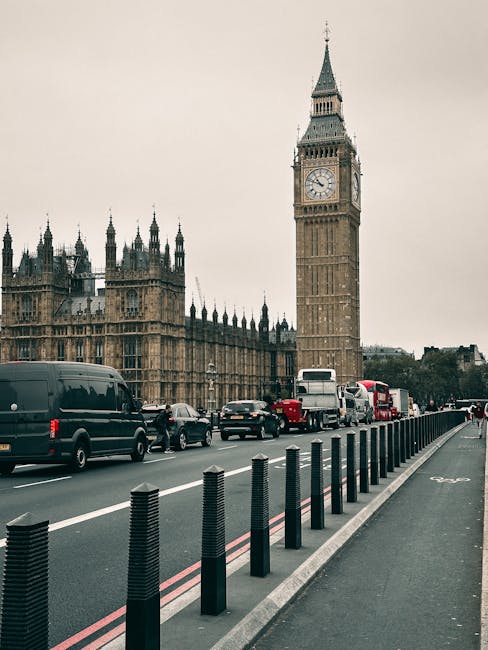 An urban scene featuring the iconic Big Ben clock tower and the Palace of Westminster in London, with several vehicles including buses, cars, and vans traveling along the road in front. The street is lined with black bollards separating the traffic lanes from the sidewalk, which is visible on the right. Overcast sky creates diffused lighting, enhancing the historic architecture and busy city atmosphere. The image emphasizes the transportation infrastructure and the classic Gothic Revival style of the buildings, with minimal visible signs of dirt or disrepair. This setting exemplifies the need for thorough surface cleaning and sanitisation, aligning with the services provided by Deep Cleaning Lambeth for efficient, same-day emergency cleaning solutions in busy urban environments.