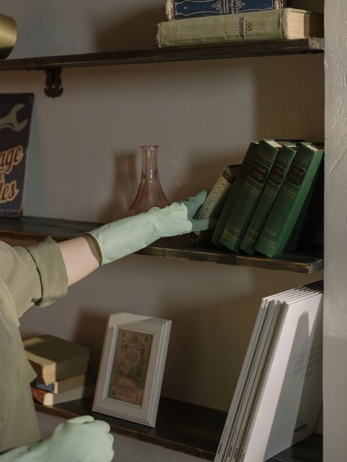 Close-up of a person's hand wearing a light green cleaning glove, reaching towards a dark wooden bookshelf with a worn edge. The shelf holds a row of green hardcover books with gold lettering on the s